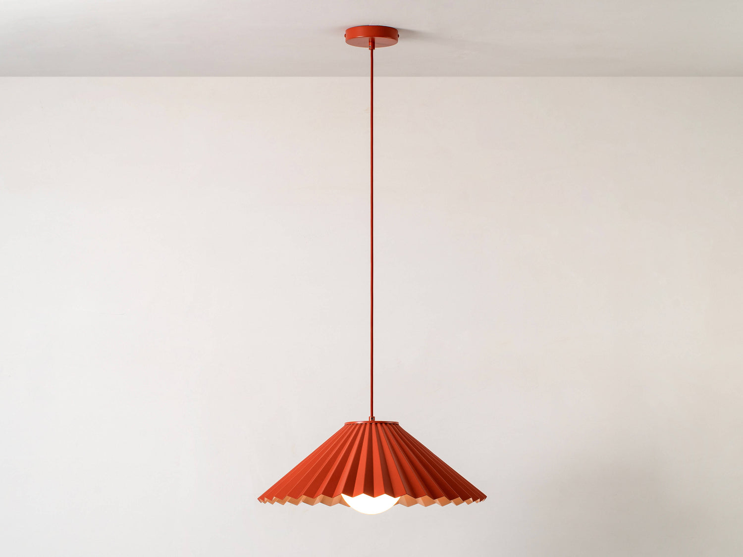 A large, fluted, pleated metal shade is suspended from a ceiling pendant. It is painted burnt orange on the outside, and pale pink on the inside. Light is on.