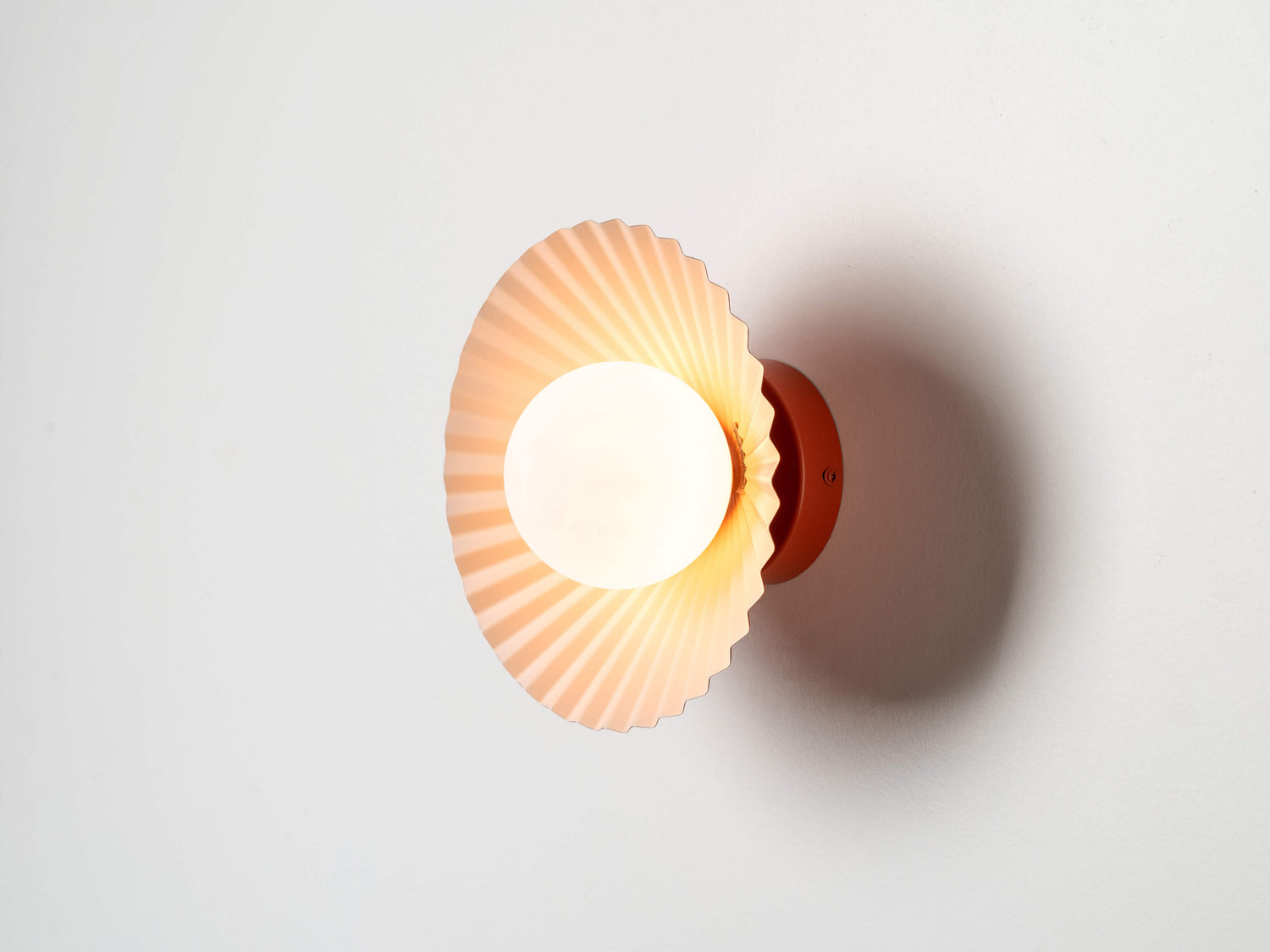 An opal glass shade is contained within fluted, pleated burnt orange coloured metal and sits close to the wall or ceiling. The inside of the pleated shade is pale pink and the outer is burnt orange. The light is on.
