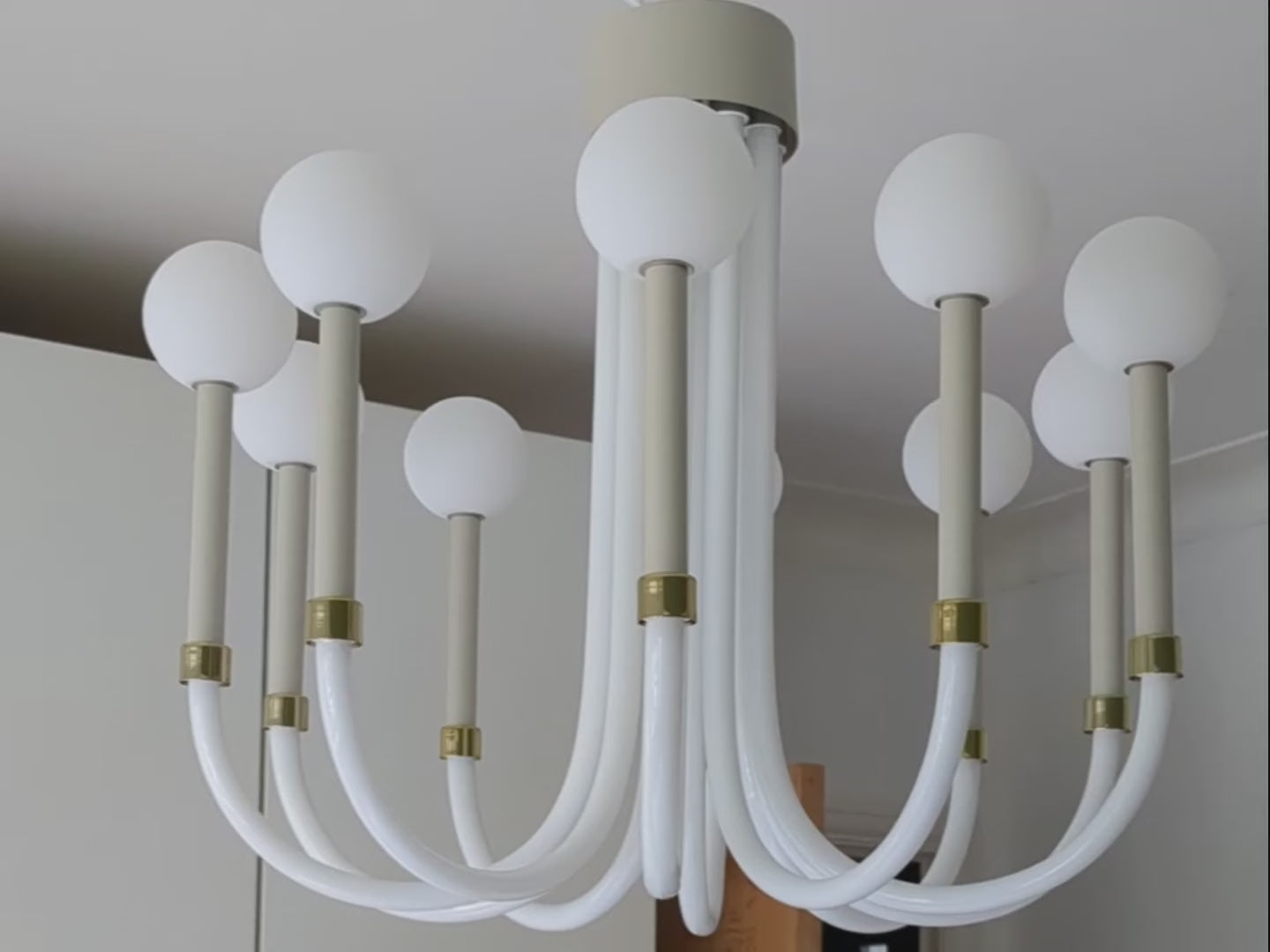 10 opaque sand-coloured glass arms are suspended from a metal ceiling fixture. The curved  sand-coloured U-shaped arms are each topped with an opal glass shade that surrounds a lamp holder. Light is shown in a few room and close ups of the light.