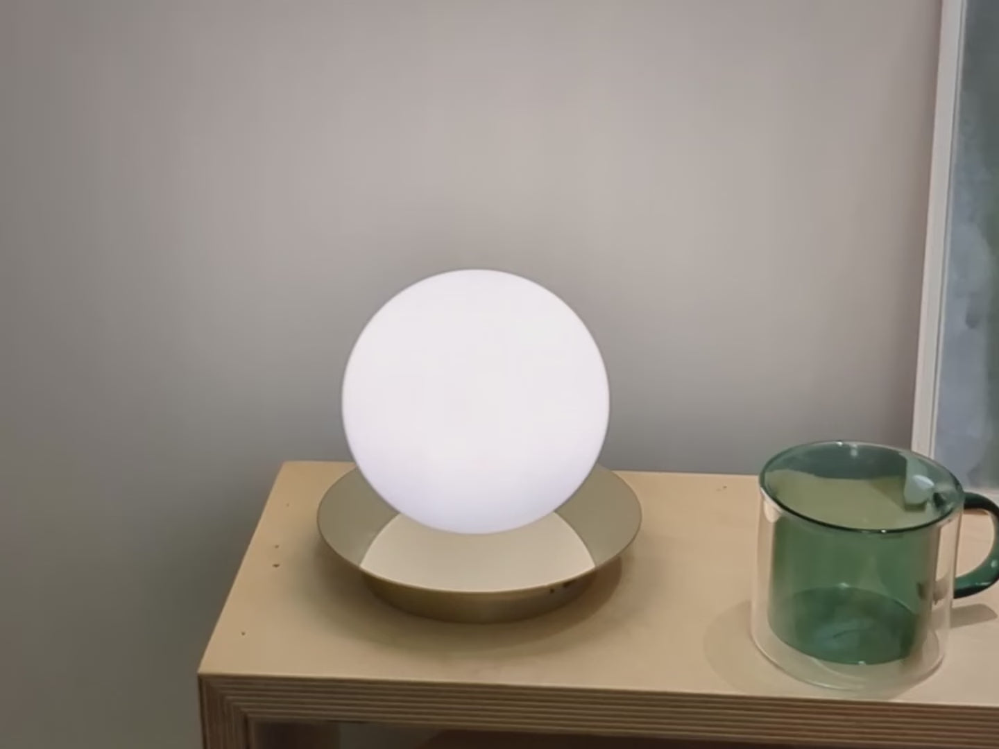 An opal glass shade sits upon a metal disk. The opal glass shade contains an integrated LED bulb that can be recharged. The lamp is portable and wireless. Light is on and on a bedside table.