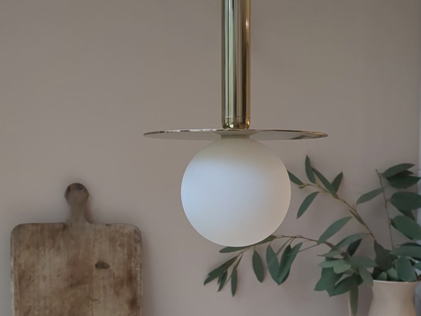 Close-up - from different angles - of a brass pendant ceiling light with a brass oval body and a horizontal brass metal plate that holds a white opal glass shade. Light is off.