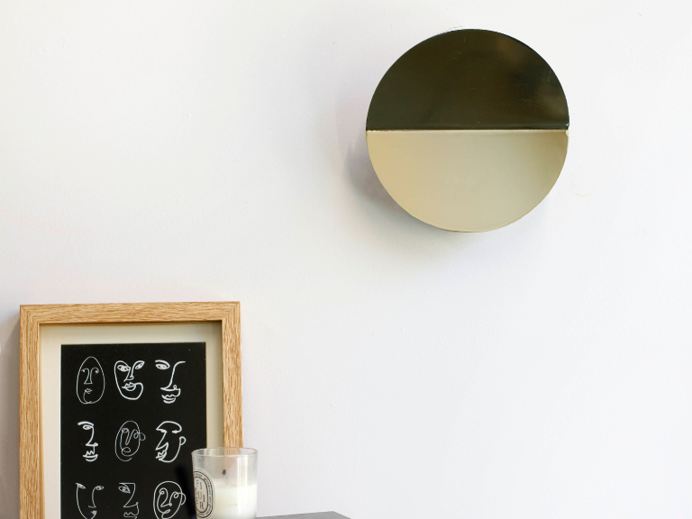 A brass circular wall light that sits flush to the wall, with a subtle fold and integrated LED bulbs installed on a white wall with a framed print and candle below.