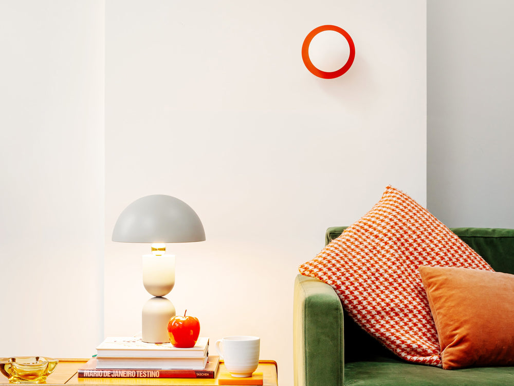 A living room with a green sofa and orange cushions with a coffee table, on which sits the mushroom dome table lamp in sand and colourful props. On the wall the opal disk wall light in orange is installed.
