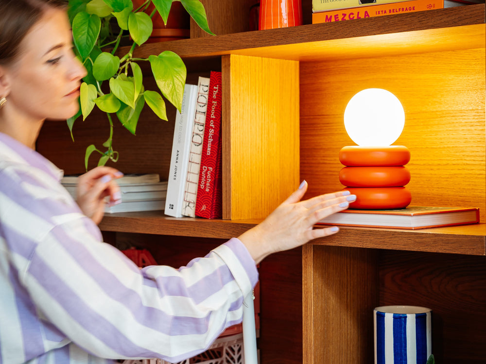 The orange rechargeable tube table lamp is positioned on wooden shelves, with other shelves decorated with cook books and colourful decor. A woman in a lilac striped shirt reaches to switch on the light - she is blurred.