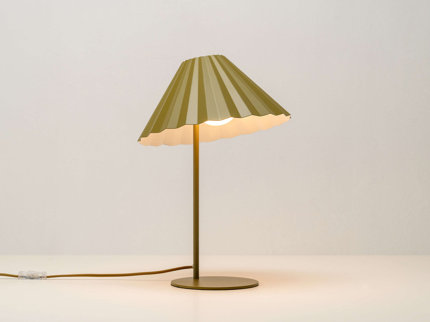 A table lamp with a fluted, pleated metal shade that can be tilted and adjusted. It is painted moss green, with a contrasting pale blue colour inside the shade. Light is on.