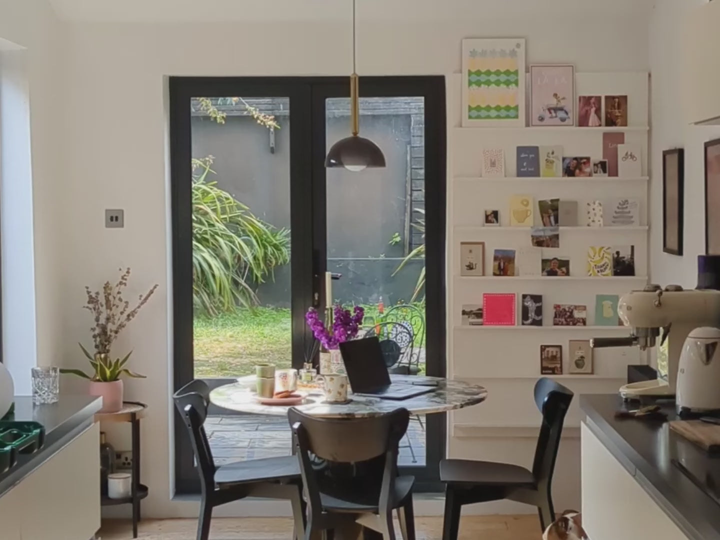 A pendant light that features a brass oval metal body attached to a charcoal grey glass dome shade which encases an opal glass shade. Set in a kitchen above a green marble dining table against big windows revealing the garden outside.