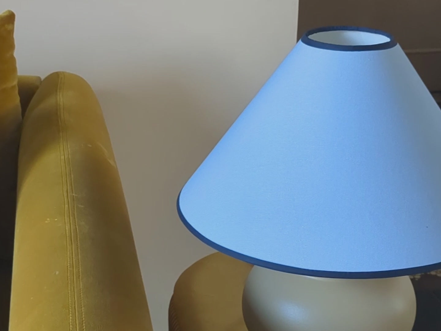 A blue plain fabric cone lamp shade with a small opening at the top (also known as a coolie shape) has a navy blue tonal-coloured trim around both edges. It has a reversible gimble so it can be used as an uplighter or downlighter.  The light is shown in a living room and is turned on by someone. 