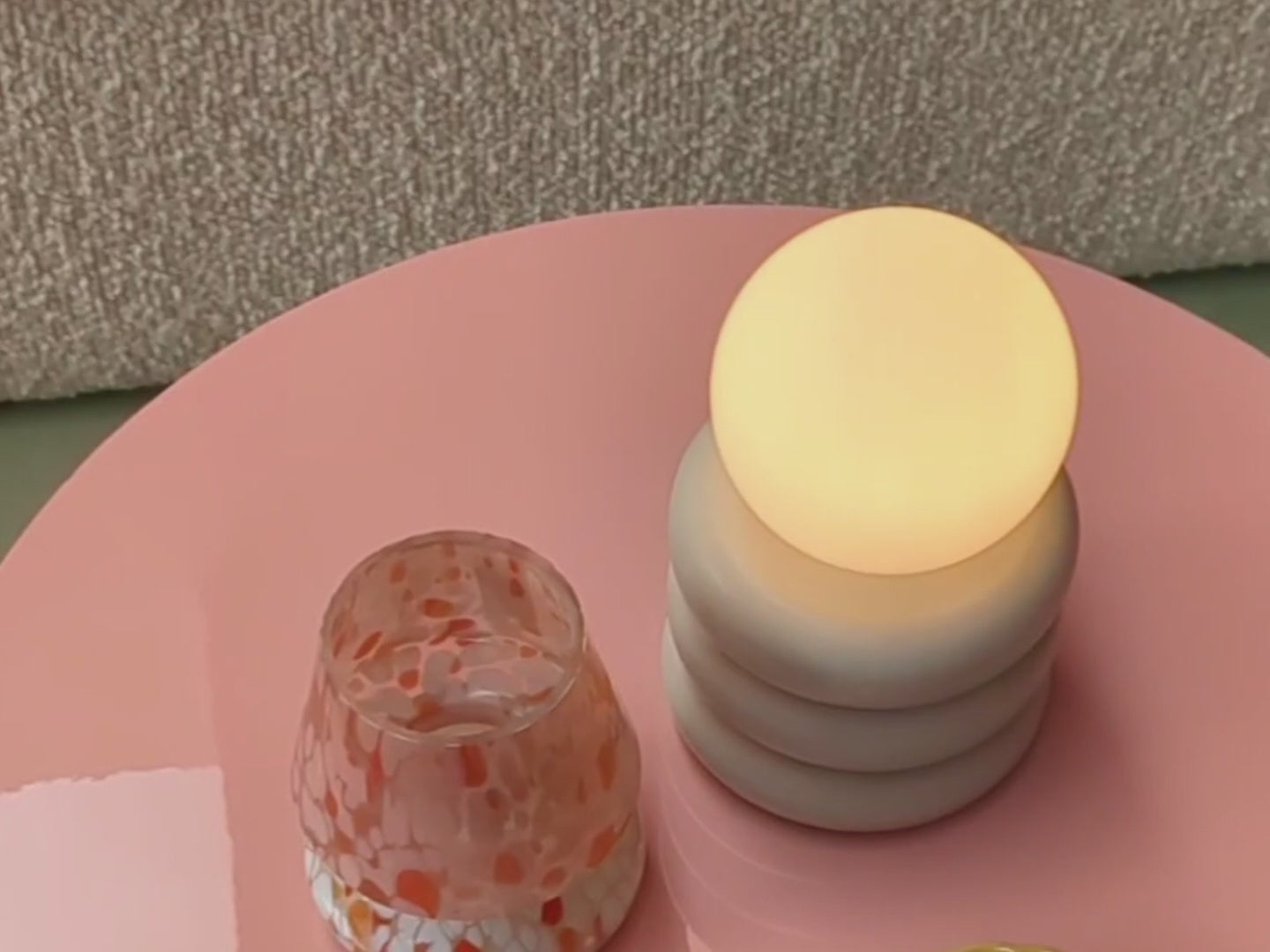 A cordless lamp consisting of three sand-coloured metal tubular discs supports an opal glass shade. The light sits on a pink table. The opal glass shade contains an integrated LED bulb that can be recharged. The lamp is portable and wireless.
