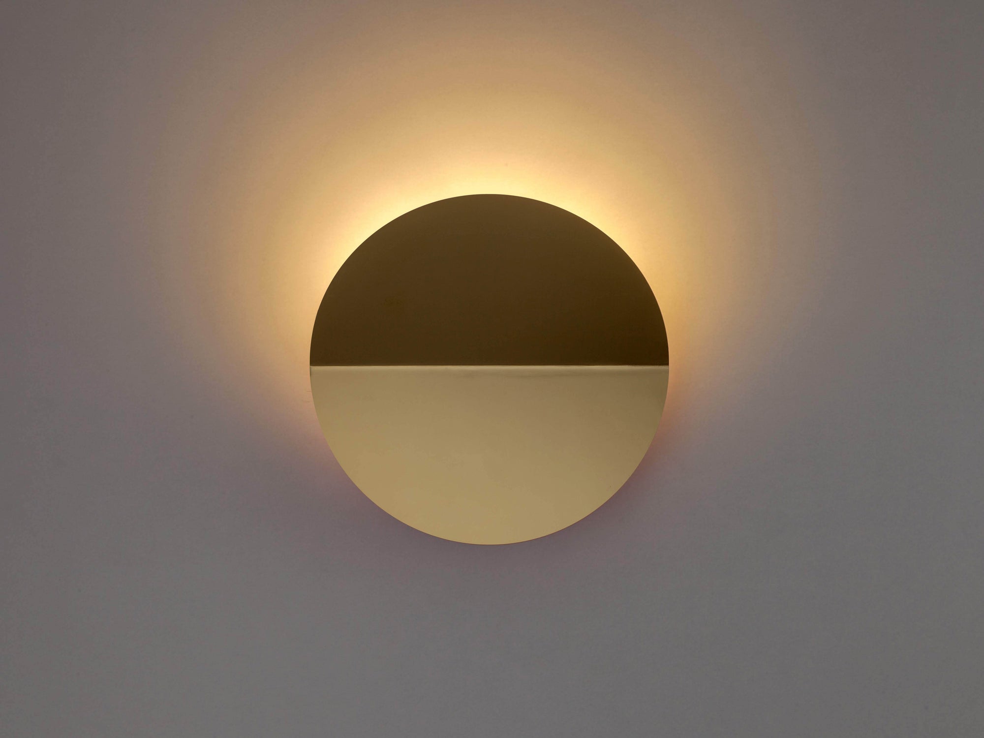 A brass circular wall light that sits flush to the wall, with a subtle fold and integrated LED bulbs. The light is on in a dark room, demonstrating how the light diffuses. 